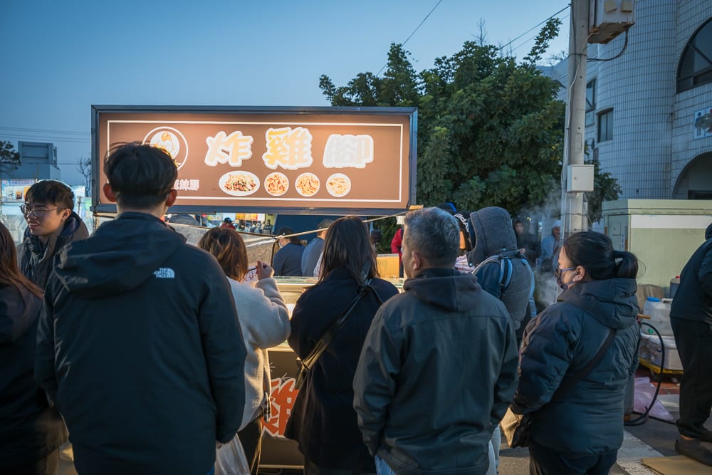苗栗後龍夜市完整攻略|週日封街市集與在地小吃指南 7 苗栗後龍夜市|街頭攤販,招牌,人群,排隊,燈箱,樹木,建築物。夜幕下路邊小吃攤前聚集多名顧客,背對鏡頭排隊等候,上