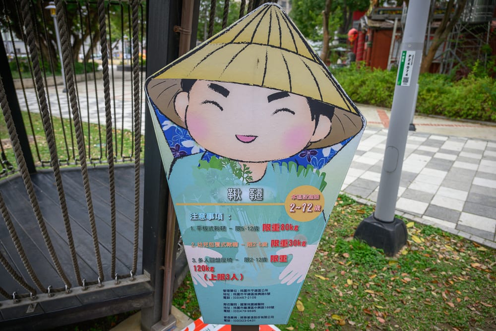 桃園民俗文化公園完整指南|兒童遊樂設施與園區玩法解析 9 桃園民俗文化公園|立牌,告示牌,農夫插畫,兒童遊樂場,繩索,地面,樹木。戶外公園設置帶斗笠農夫插畫的告示牌,標示遊具使
