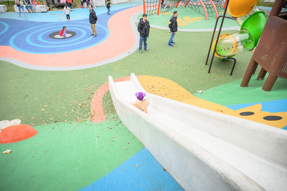 桃園民俗文化公園完整指南|兒童遊樂設施與園區玩法解析 11 桃園民俗文化公園|溜滑梯、兒童遊樂場、彩色地坪、大人、小孩,俯瞰視角捕捉戶外公園設施與活動人群,地表散落落葉,綠色、橘