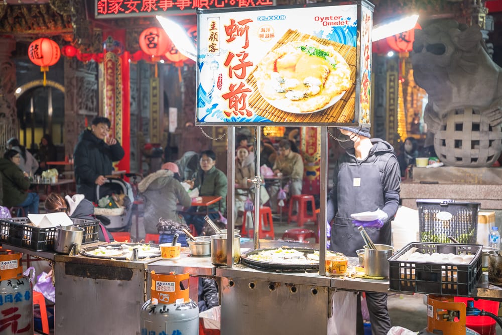 苗栗後龍夜市完整攻略|週日封街市集與在地小吃指南 6 苗栗後龍夜市|夜市攤販、蚵仔煎招牌、平底鍋、瓦斯桶、石獅雕像、紅燈籠、人群。攤販前方的招牌寫著蚵仔煎與Oyster