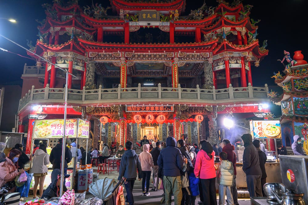 苗栗後龍夜市完整攻略|週日封街市集與在地小吃指南 9 苗栗後龍夜市|寺廟,夜市,人群,攤販,紅燈籠,招牌,夜間燈光。夜間寺廟前聚集人潮,紅柱飛檐建築下擺設多個小吃攤位,