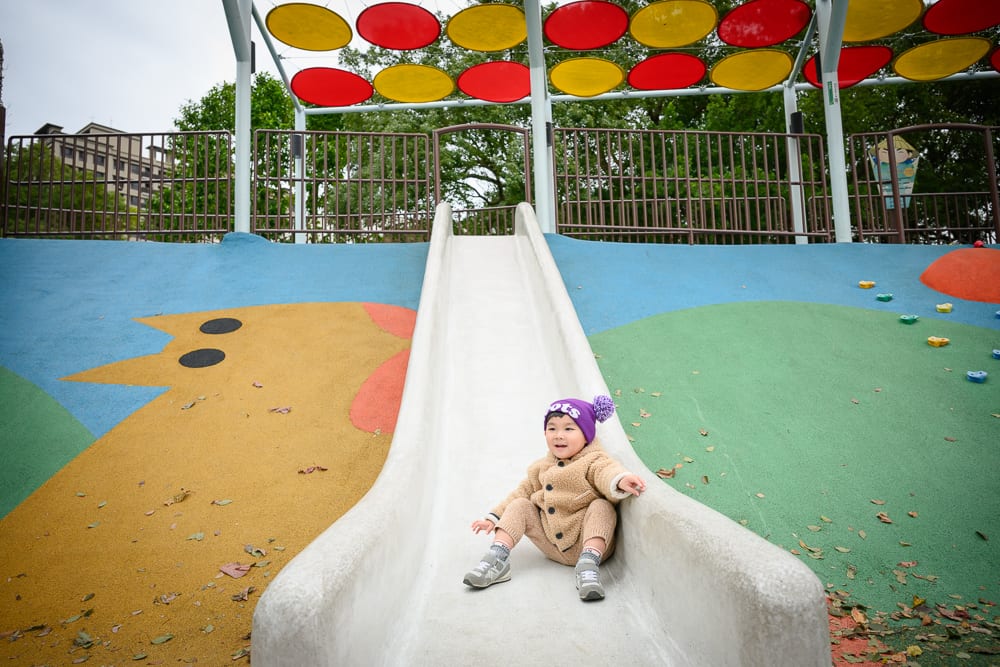 桃園民俗文化公園完整指南|兒童遊樂設施與園區玩法解析 12 桃園民俗文化公園|兒童溜滑梯,公園,幼童,彩色地墊,遮陽棚。一名穿著棕色外套與紫色毛帽的幼童坐在溜滑梯底部。兩側地面繪
