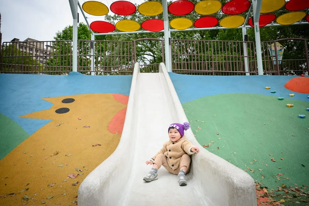 桃園民俗文化公園｜兒童溜滑梯，公園，幼童，彩色地墊，遮陽棚。一名穿著棕色外套與紫色毛帽的幼童坐在溜滑梯底部。兩側地面繪