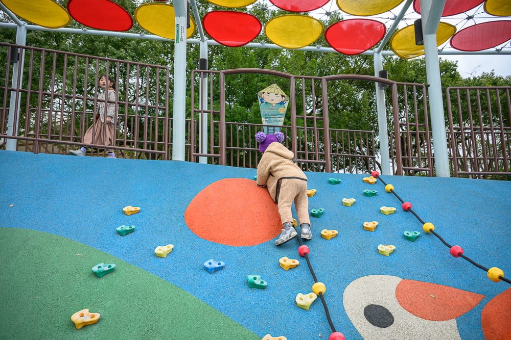 桃園民俗文化公園完整指南|兒童遊樂設施與園區玩法解析 7 桃園民俗文化公園|兒童遊戲場,攀爬牆,小孩,金屬欄杆,遮陽棚,綠樹。穿著棕色毛茸外套與紫色帽子的孩童正在攀爬藍色斜坡,