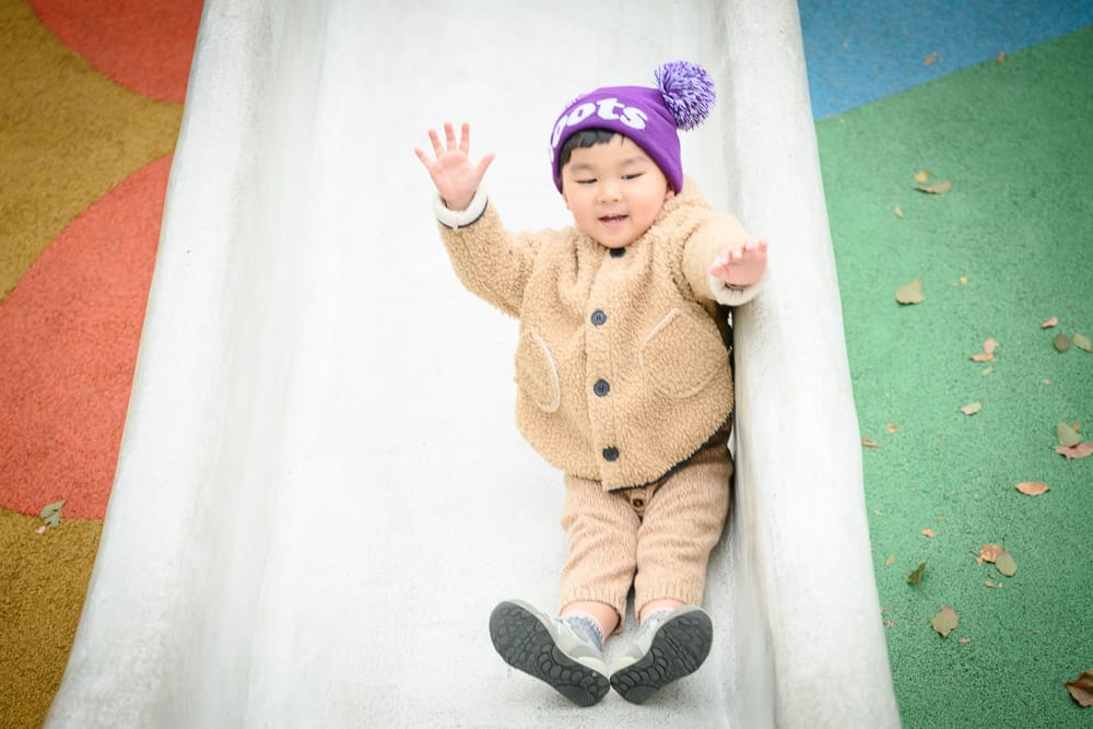 桃園民俗文化公園完整指南|兒童遊樂設施與園區玩法解析 10 桃園民俗文化公園|兒童、溜滑梯、毛帽、外套、鞋子、遊樂場,戴著紫色球帽的小孩坐在白色滑梯上,雙手張開,穿著米色羊毛外套
