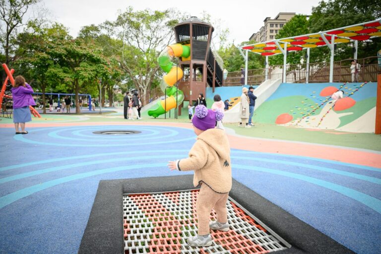 桃園民俗文化公園完整指南|兒童遊樂設施與園區玩法解析 19 桃園民俗文化公園|公園、彈跳床、溜滑梯、攀爬牆、兒童、樹木、建築。幼童穿著卡其色毛绒外套與紫色毛帽,站在嵌入地面的網狀