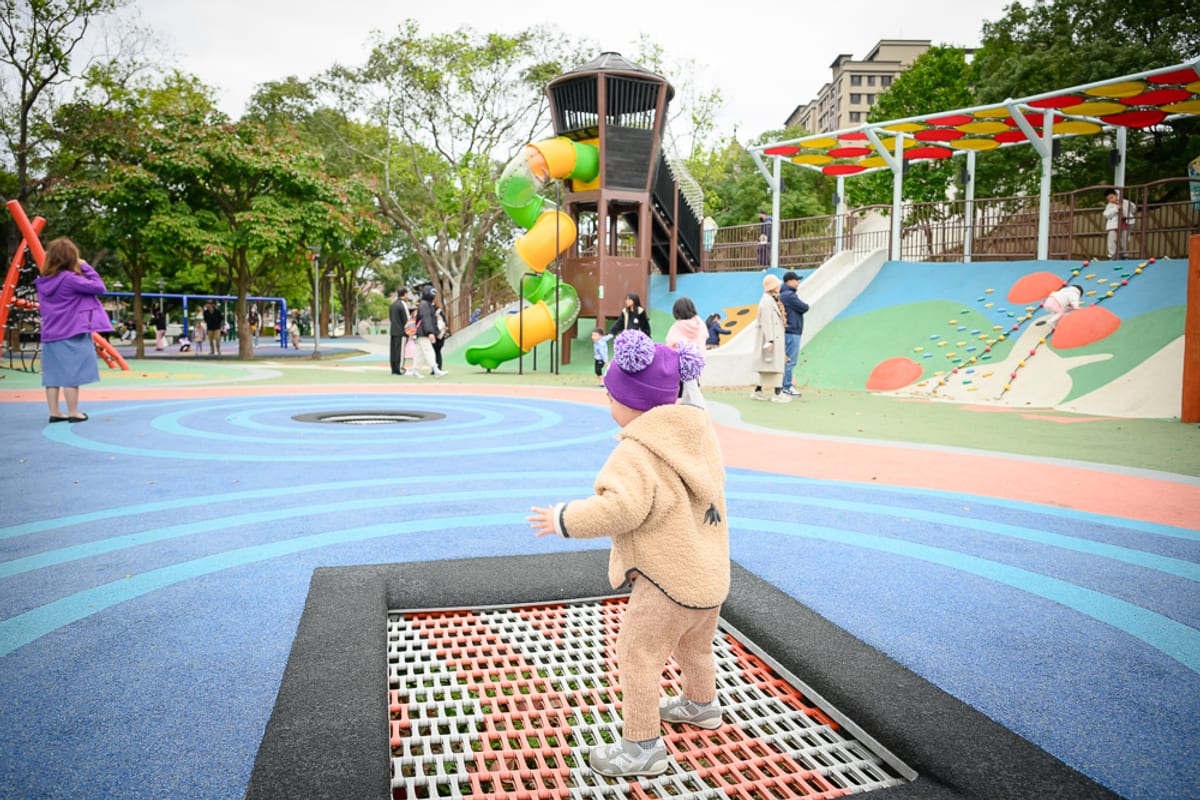 桃園民俗文化公園完整指南|兒童遊樂設施與園區玩法解析 1 桃園民俗文化公園|公園、彈跳床、溜滑梯、攀爬牆、兒童、樹木、建築。幼童穿著卡其色毛绒外套與紫色毛帽,站在嵌入地面的網狀