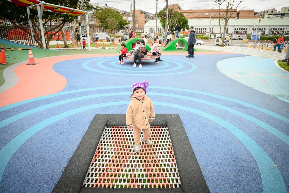 桃園民俗文化公園完整指南|兒童遊樂設施與園區玩法解析 13 桃園民俗文化公園|戶外公園,彈跳床,小孩,彩色地墊,旋轉遊具,遠處建築,陰天光線