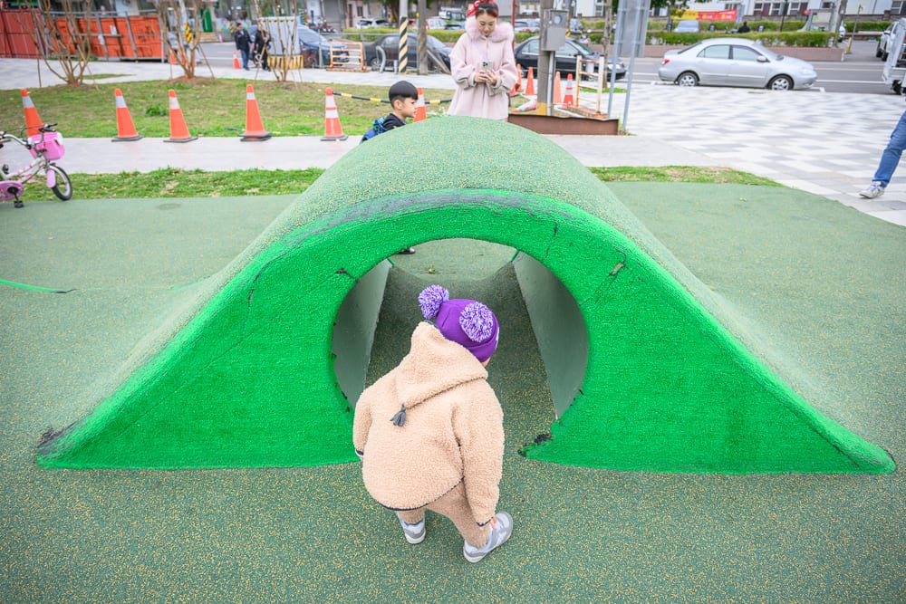 桃園民俗文化公園完整指南|兒童遊樂設施與園區玩法解析 8 桃園民俗文化公園|兒童遊戲場,綠色人工草皮山丘,爬行隧道,穿著毛茸茸外套與紫色毛球帽的孩童,後方站立成年人,背景街道,