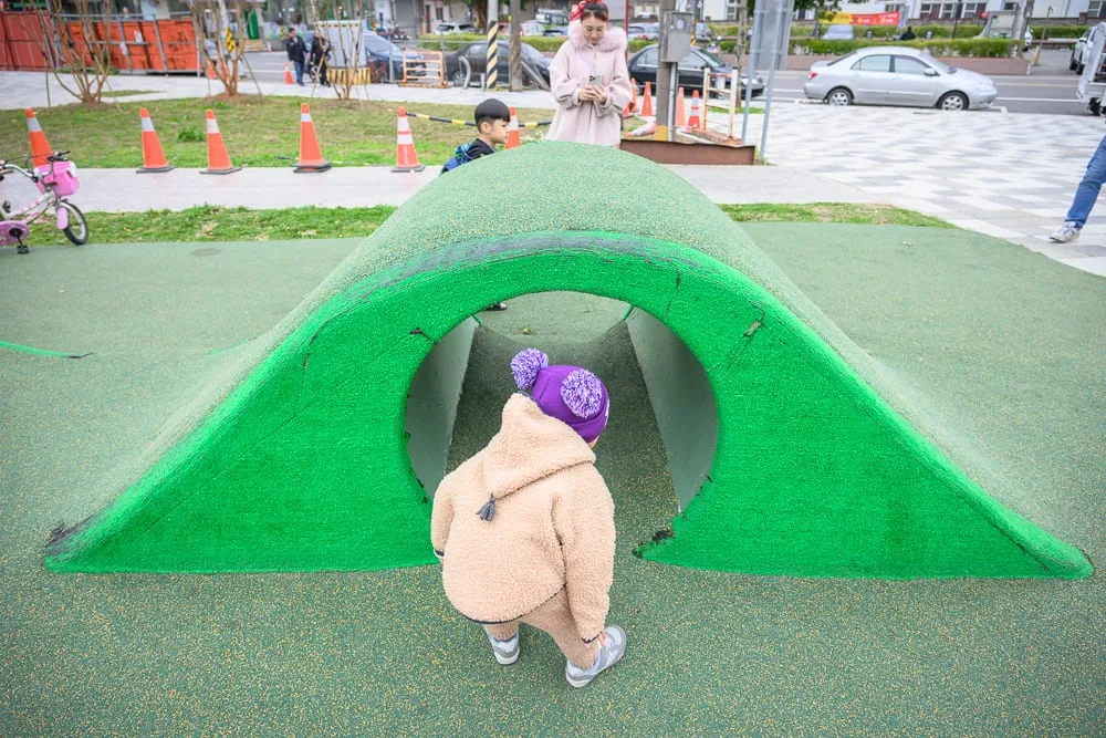 桃園民俗文化公園｜兒童遊戲場，綠色人工草皮山丘，爬行隧道，穿著毛茸茸外套與紫色毛球帽的孩童，後方站立成年人，背景街道，