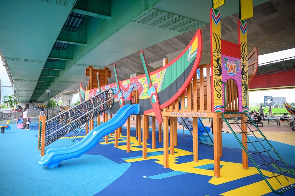 宜蘭礁溪二龍之心公園｜橋下空間，龍舟造型遊具，溜滑梯，攀爬網，藍黃色地墊，木質支架，行人，遠方建築。