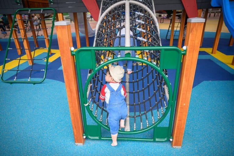 宜蘭礁溪二龍之心公園｜兒童遊樂場，幼童，吊橋，繩網爬架，木柱，藍色塑膠地墊。戴帽子的幼童穿著吊帶褲，正在攀爬圓形網狀隧道，
