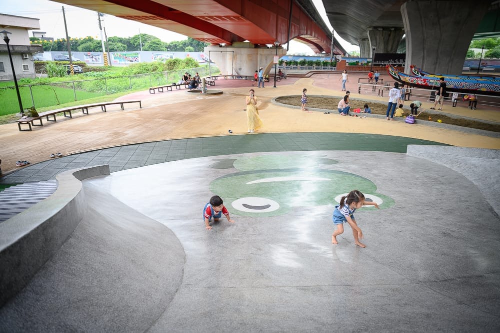 宜蘭礁溪二龍之心公園｜高架橋下廣場、溜滑梯、青蛙圖案、兒童、沙坑、龍舟模型，地面鋪設水泥與地墊，多位大人小孩在陰影下活動，