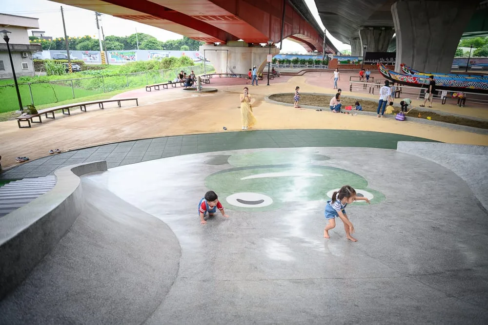 宜蘭礁溪二龍之心公園｜高架橋下廣場、溜滑梯、青蛙圖案、兒童、沙坑、龍舟模型，地面鋪設水泥與地墊，多位大人小孩在陰影下活動，