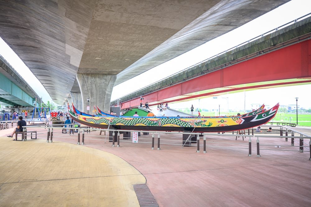 宜蘭礁溪二龍之心公園｜高架橋下廣場展示彩繪龍舟，地面鋪設紅磚，遠處有遊具與綠地，橋墩與紅色鋼梁交錯，民眾三三兩兩散步休憩。
