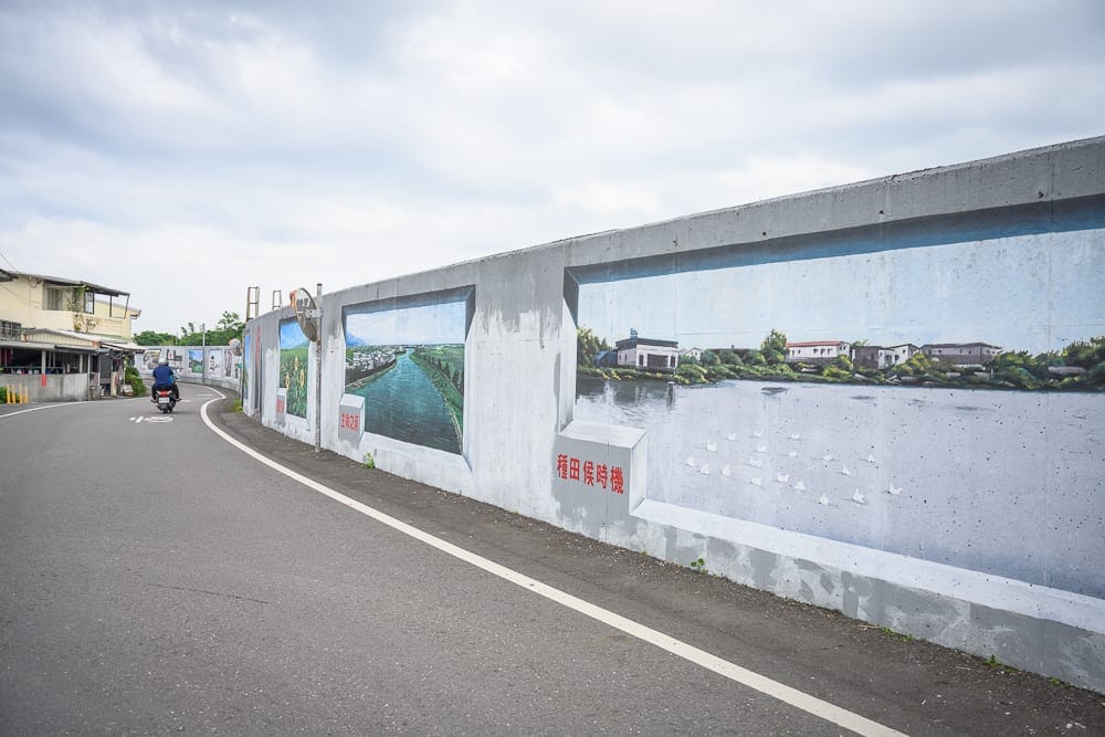 宜蘭礁溪二龍之心公園｜彎曲的柏油道路旁，高聳的混凝土堤防繪有多幅風景壁畫與紅字標語，遠處有騎機車的人影，陰天雲層籠罩的天空