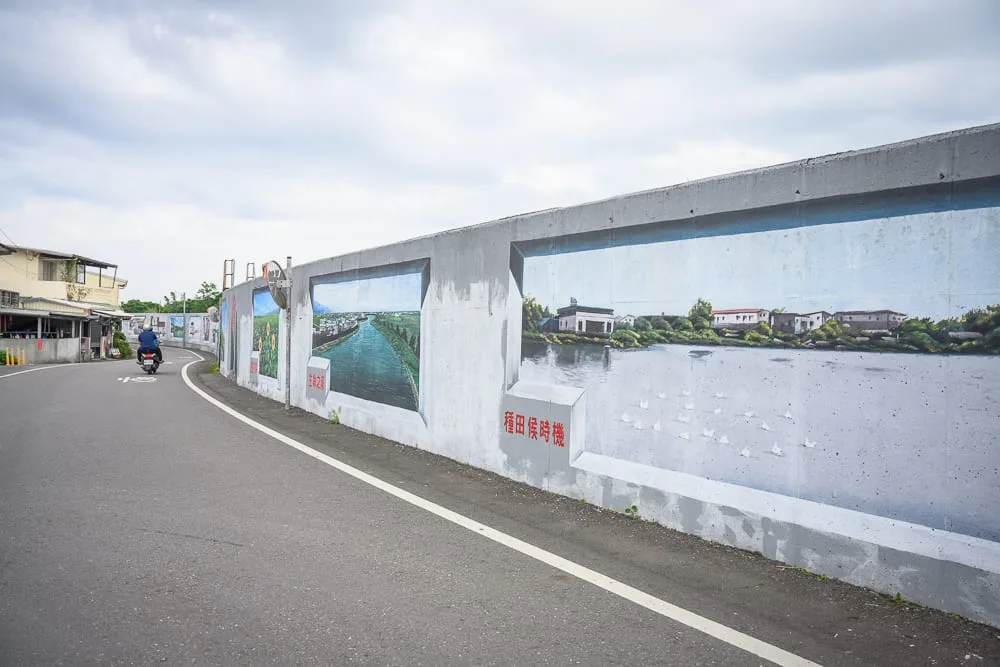 宜蘭礁溪二龍之心公園｜彎曲的柏油道路旁，高聳的混凝土堤防繪有多幅風景壁畫與紅字標語，遠處有騎機車的人影，陰天雲層籠罩的天空