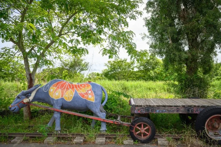 嘉義板頭車站公園完整指南|交趾陶剪黏藝術與五分車鐵道玩法 14 嘉義板頭車站公園|戶外,草地,鐵道,水牛雕塑,木板車,大樹。藍灰色水牛身側有橙色馬賽克蝶紋,背部連接雙輪板車,停在綠草