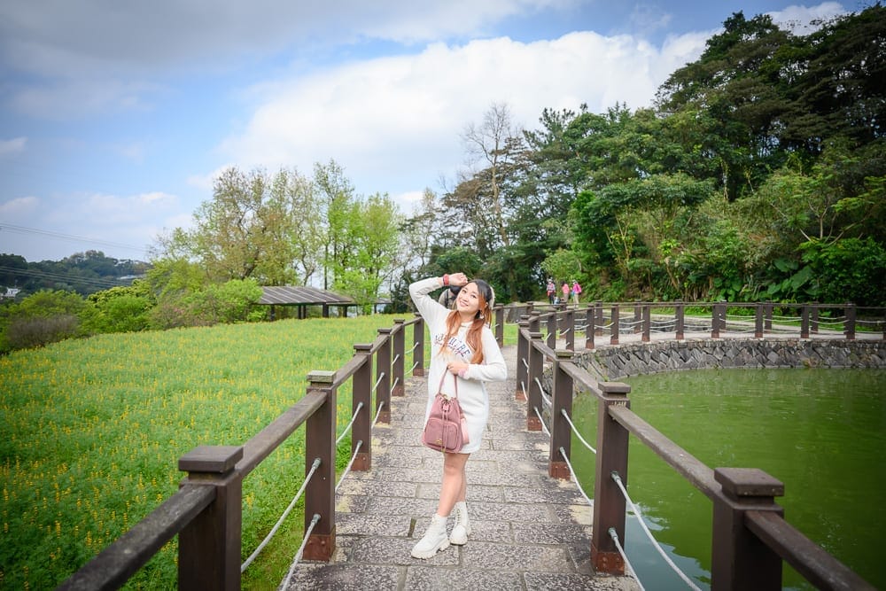 台北樟湖步道魯冰花｜女性、木棧道、水池、草地、樹林、藍天白雲。女子背著粉色背包站立在石板木棧道中央，右手托腮微笑，一側是