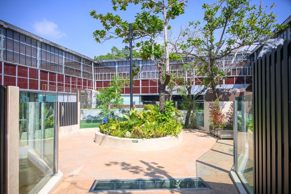 台南頑皮世界｜中庭、樹木、植栽、建築、玻璃帷幕、藍天。露天中庭中央為圓形花圃，周圍環繞雙層廠房式建築，牆面帶有紅色