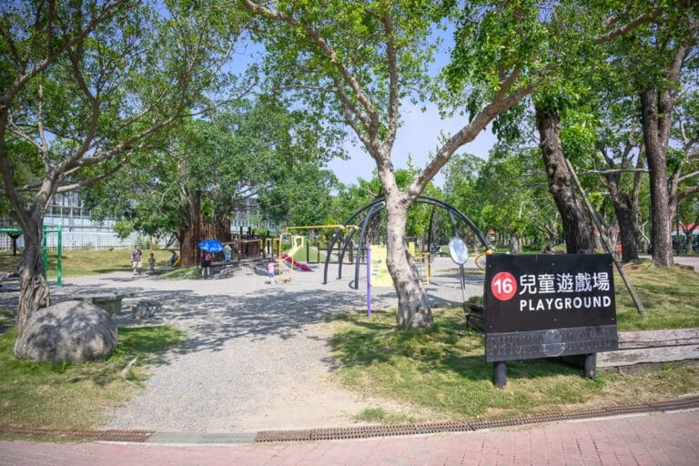 台南頑皮世界野生動物園完整攻略|動物餵食、水樂園與親子必玩設施 9 台南頑皮世界|綠樹、草地、兒童遊戲場、指標、攀爬架、溜滑梯、大石塊、行人。露天公園中設有金屬攀爬架與木造遊具,右側