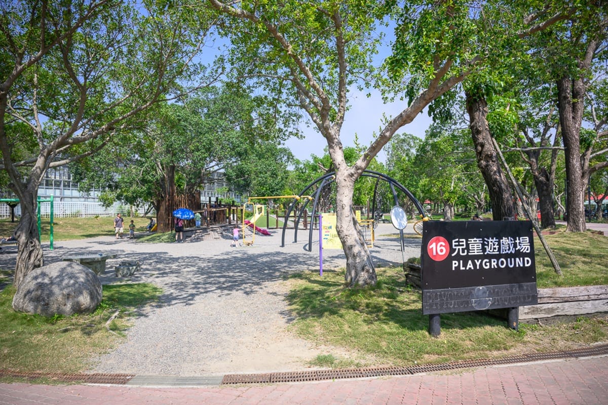 台南頑皮世界｜綠樹、草地、兒童遊戲場、指標、攀爬架、溜滑梯、大石塊、行人。露天公園中設有金屬攀爬架與木造遊具，右側