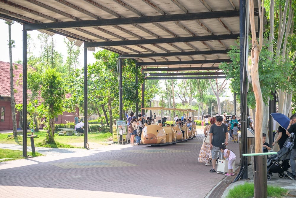 台南頑皮世界｜戶外走廊，鐵皮屋頂，磚造地面，遊園車，水豚造型車頭，人群，綠樹，草皮，遮陽傘，嬰兒車
