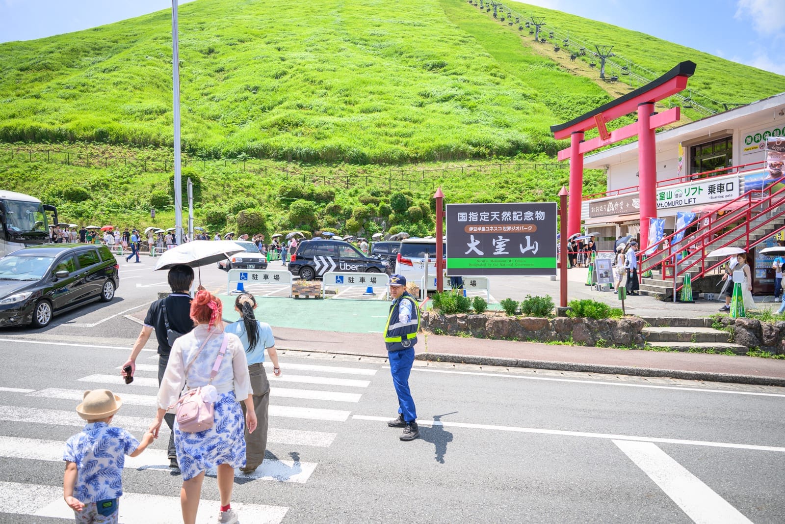 日本靜岡大室山纜車完整攻略|死火山口山頂遠眺富士山玩法解析 8 日本靜岡-大室山纜車|行人、斑馬線、導引警察、山脈、紅色鳥居、汽車,前方綠草如茵的大山背景下,遊客走在戶外馬路的行人穿越道