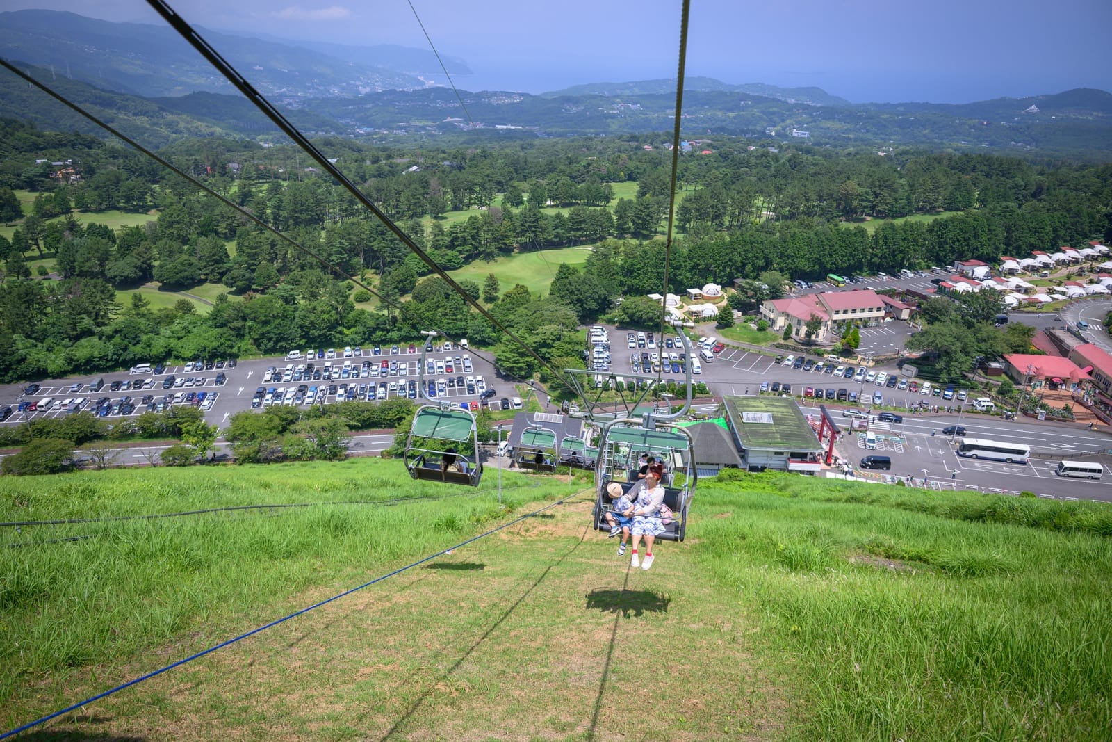 日本靜岡大室山纜車完整攻略|死火山口山頂遠眺富士山玩法解析 12 日本靜岡-大室山纜車|纜車、纜繩、草坡、樹林、停車場、遠山、建築,空中吊椅載著乘客下降,下方鋪滿綠草斜坡與密林,遠方海景與