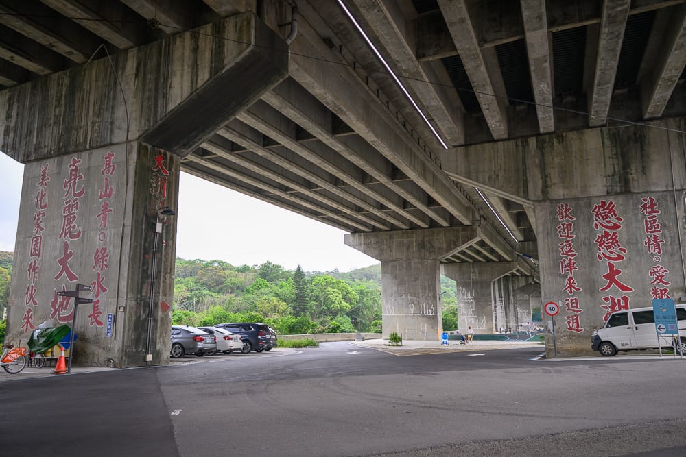 新竹風火輪公園｜高架橋、橋墩、柏油路面、汽車、宣傳標語、遠方山林。水泥橋墩上漆有紅色大字，橋下為寬闊的道路與停車空間