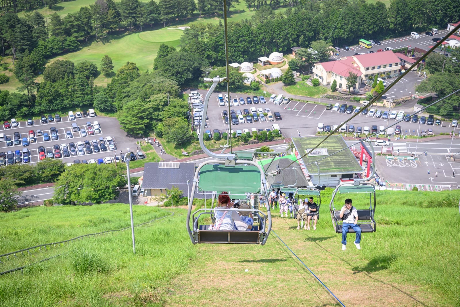 日本靜岡大室山纜車完整攻略|死火山口山頂遠眺富士山玩法解析 13 日本靜岡-大室山纜車|纜車、草坡、停車場、建築、樹林、車輛,遊客搭乘懸掛式纜車下降,俯瞰下方綠地、建築與停滿車輛的停車場。