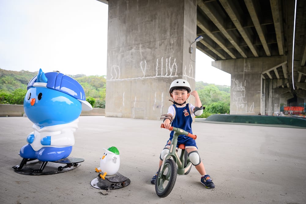 新竹風火輪公園｜兒童、滑步車、公仔、高架橋下、水泥地。戴著安全帽的小孩坐在滑步車上，旁側立著藍色與白色的大型卡通公仔