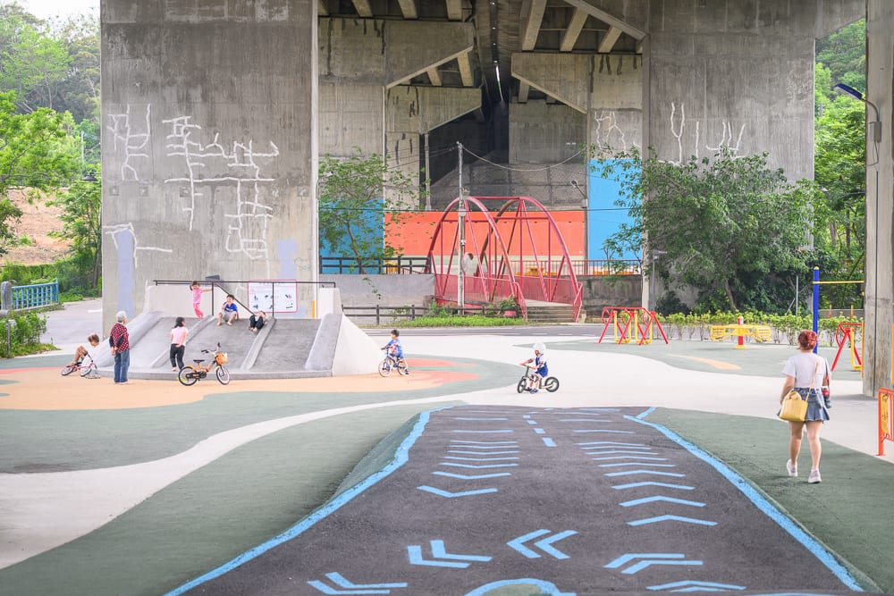 新竹風火輪公園｜高架橋下空間、溜滑梯、單車、兒童、行人、紅色拱橋、水泥柱。水泥結構下方設有彩色遊樂設施與自行車道，孩