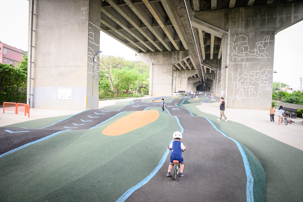 新竹風火輪公園｜橋下空間，高架橋墩，波浪形車道，兒童，平衡車，塗裝跑道，行人。幼童戴白色安全帽騎乘平衡車，前方為起伏