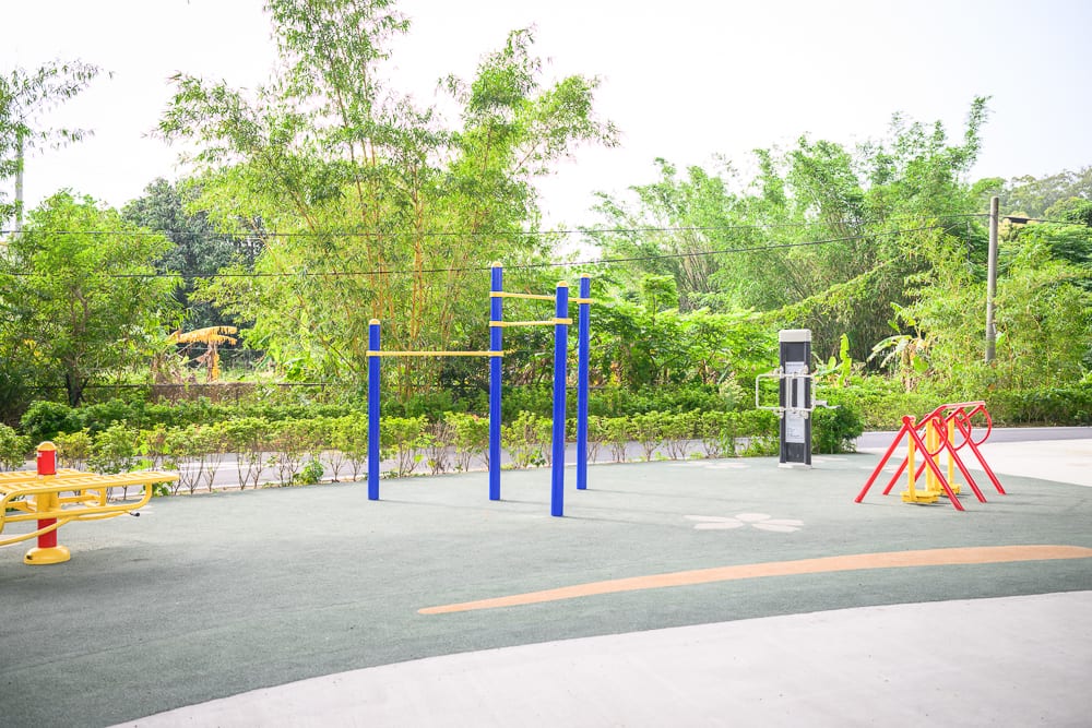 新竹風火輪公園｜戶外健身場，單槓，訓練器材，地面，灌木叢，樹木。空曠的露天場地鋪設灰色軟墊，設有藍黃相間的單槓與紅色