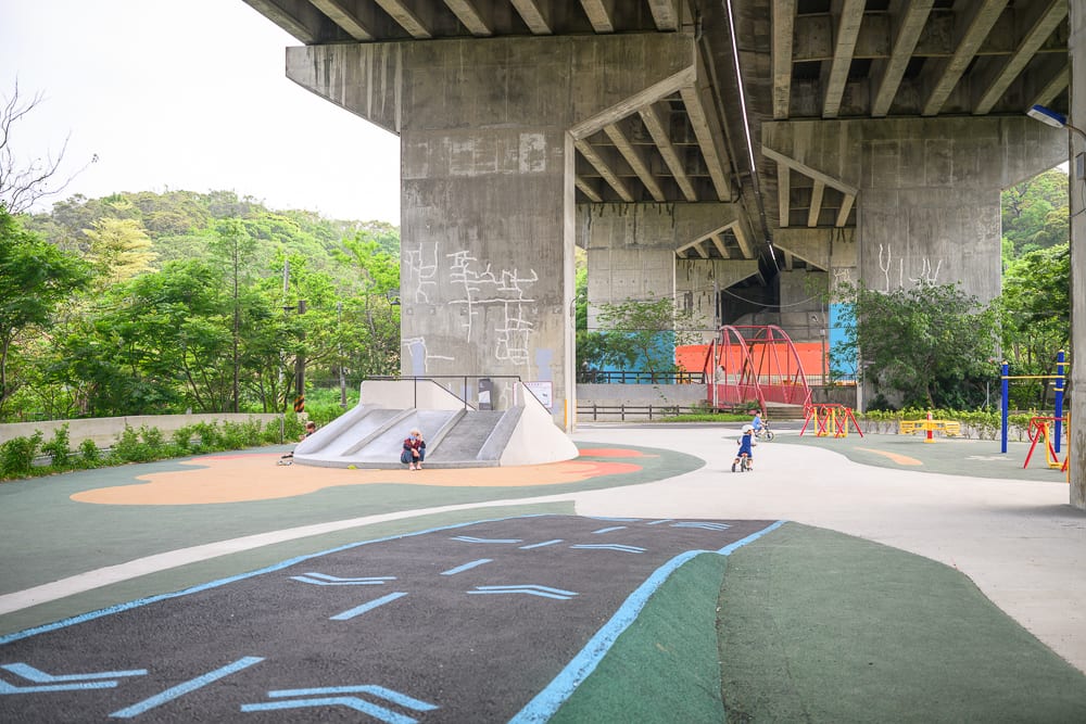 新竹風火輪公園｜高架橋、水泥橋墩、溜滑梯、彩色跑道、兒童、紅橋、樹林，水泥地面的戶外遊樂場，遠處有綠意山坡，結構錯落