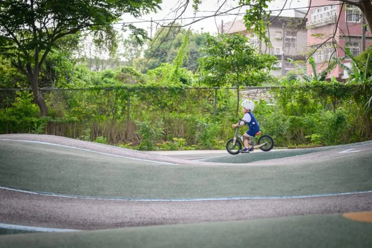 新竹風火輪公園完整指南|波浪跑道與親子設施玩法解析 5 新竹風火輪公園|戴著白色安全帽的幼童,騎著無踏板平衡車,行駛在綠色波浪狀的戶外PU跑道上。背景為茂密樹叢、金屬圍籬與