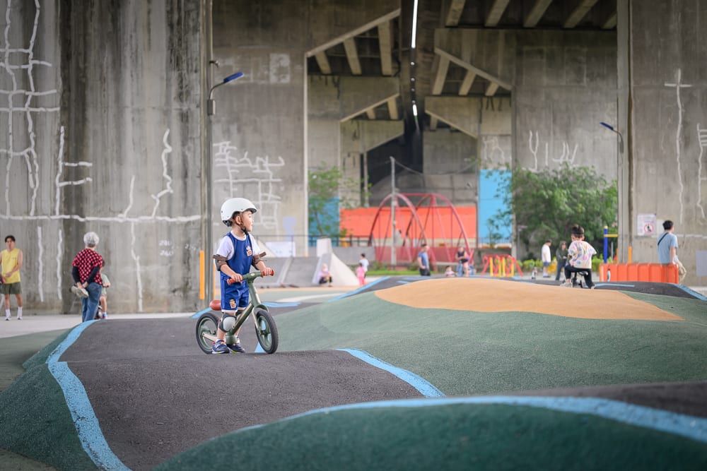 新竹風火輪公園｜高架橋下，滑步車，兒童賽道，水泥柱，公園。戴安全帽的男童跨坐綠色滑步車，停在綠黑相間的波浪起伏跑道上