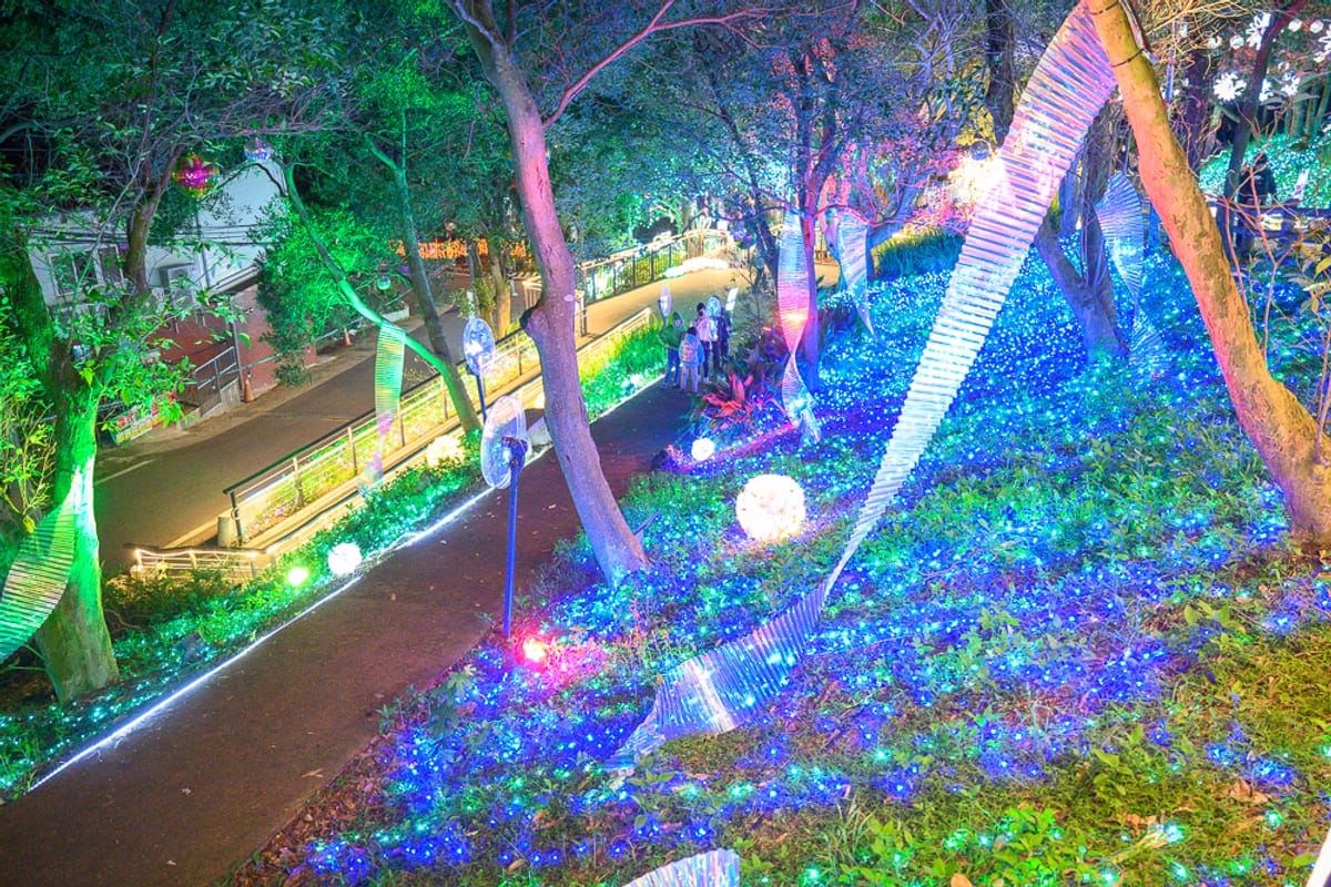 桃園大古山步道夏日繽紛派對完整攻略|夜間燈海步道玩法解析 1 桃園大古山步道夏日繽紛派對|夜間斜坡佈滿藍綠色LED燈海,樹幹間垂掛白色波浪狀燈條與球形燈飾,左側為柏油路面街道與護欄。