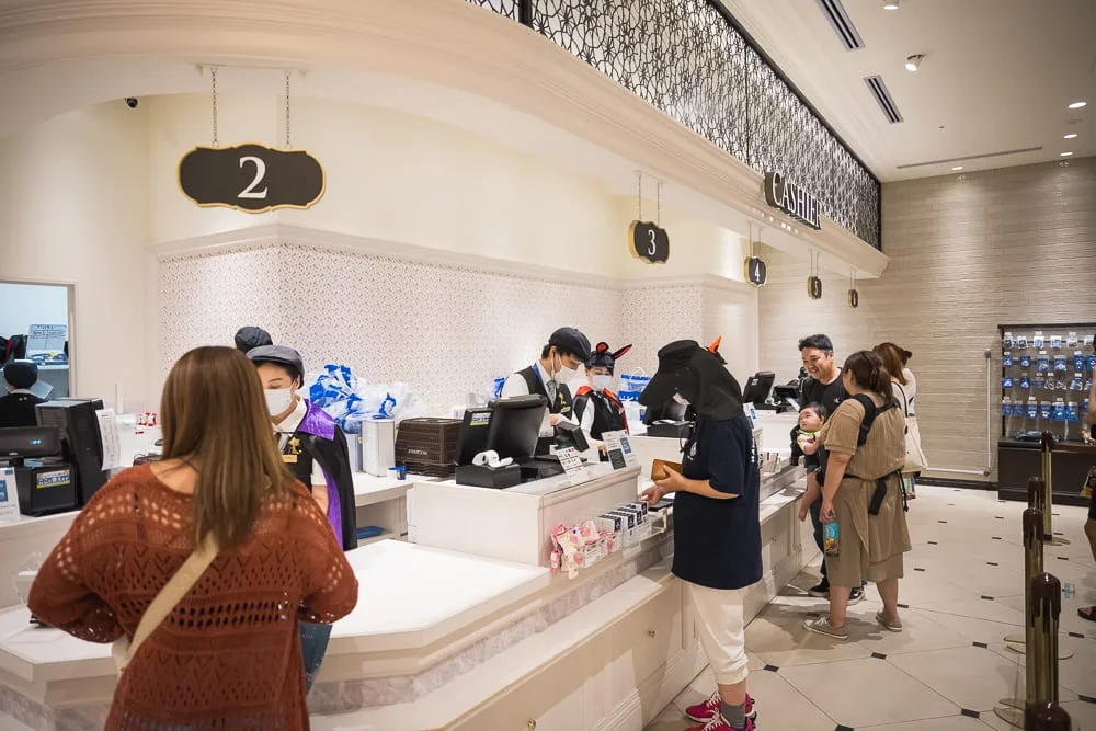 日本札幌-白色戀人觀光工廠｜室內、櫃檯、收銀員、顧客、告示牌。白色櫃檯後方站著戴口罩的收銀員，前方排隊結帳的顧客各自站立，上方懸