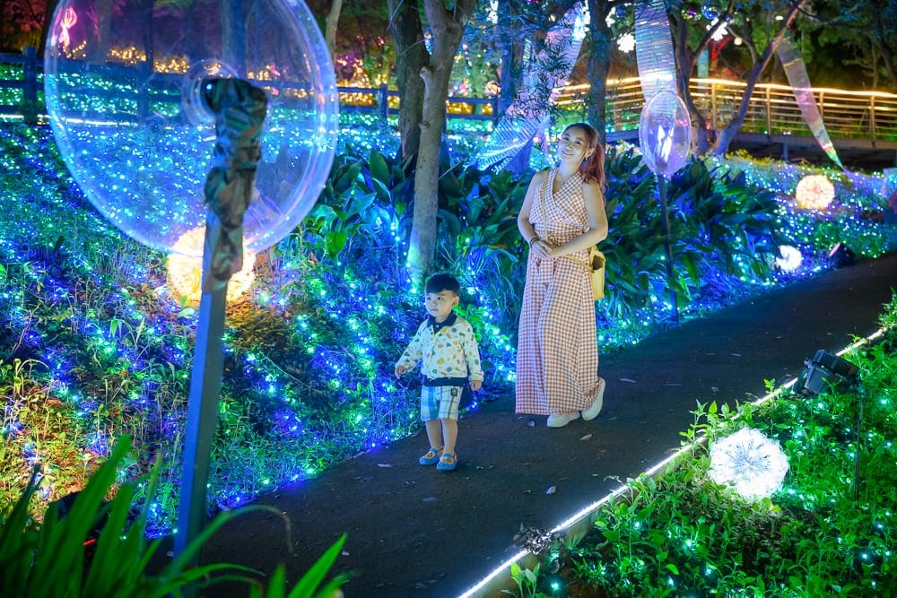 桃園大古山步道夏日繽紛派對完整攻略|夜間燈海步道玩法解析 11 桃園大古山步道夏日繽紛派對|戶外夜間步道,穿格子洋裝的女子與小男孩站立路旁,草叢覆蓋藍綠色燈串,樹林,發光球體,透明裝飾。