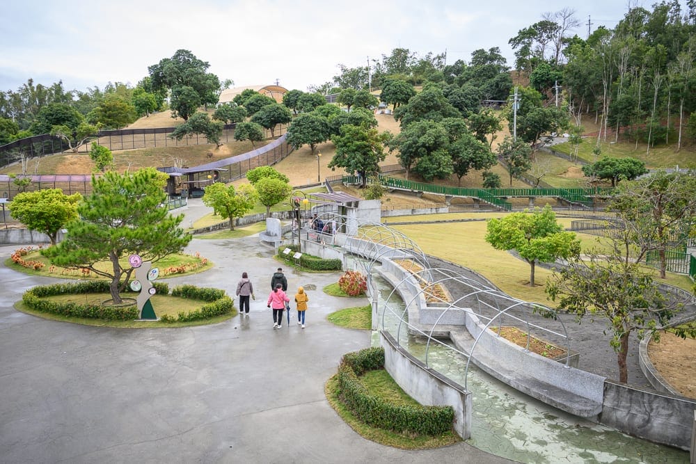 南投九九峰動物樂園完整攻略|鸚鵡互動與牧場動物玩法解析 12 南投九九峰動物園|戶外公園、水泥步道、綠樹、草皮、行人、弧形造景、遠方山丘,灰白陰天光線下,四人漫步於廣闊園區。