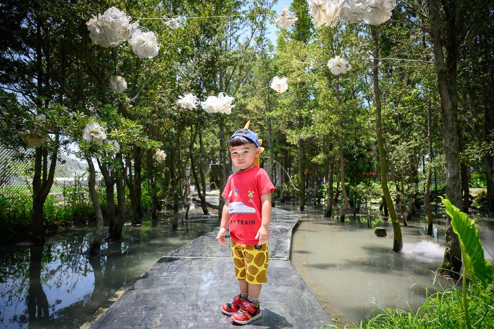 台中喜鵲方舟｜樹林、水域、走道、小孩，林間架設白色紙球裝飾，男孩身穿紅衣與黃花紋短褲，站在積水樹林中的黑色長型步道