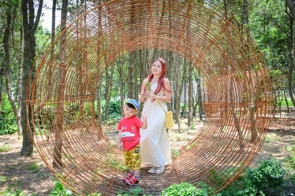 台中喜鵲方舟 fairyLand 完整攻略|梅花鹿互動與親子動物體驗 10 台中喜鵲方舟|樹林、鋼筋網圓筒裝置藝術、女性、男童、黃色斜背包、紅衣、白長裙,背景是綠樹與泥地。