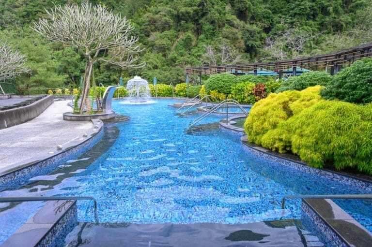 苗栗南庄雲水渡假｜泳池，噴泉，綠樹，山壁，階梯，扶手，藍色磁磚。露天溫泉池中噴水柱湧動，池邊修剪整齊的黃綠灌木叢延伸至