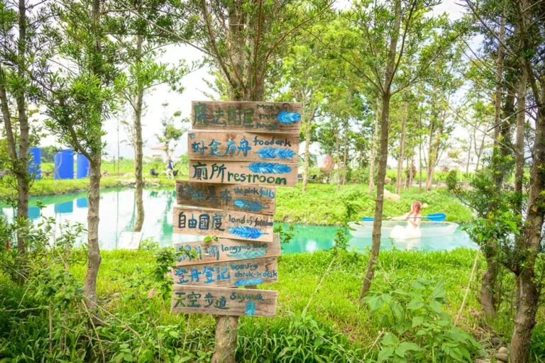 台中喜鵲方舟｜木頭指標牌、樹林、水池、草地、小船、泛舟女子。多層次的綠意中立著一根豎有多塊木板的手作指標，牌面上寫
