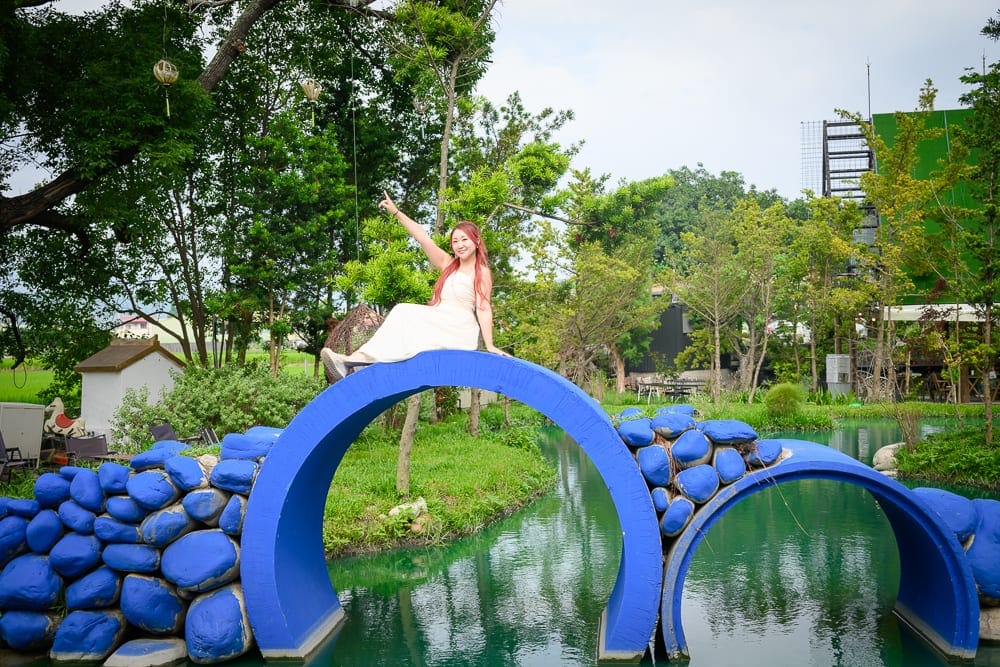 台中喜鵲方舟｜女子、藍色半圓石橋、河流、綠樹、草地，穿白裙的長髮女子坐在橋頂舉手微笑，背景有白色小屋與綠色植栽，水