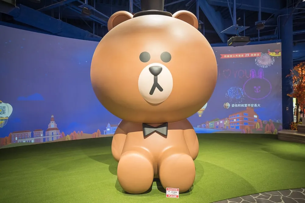 台中熊大的麗寶小鎮完整攻略|LINE FRIENDS 七大互動主題、門票與 50 分鐘玩法 13 台中麗寶樂園熊大主題|大型熊公仔模型,室內展場,草皮,黑色禮帽,黑色領結,弧形投影螢幕,藍色鋼構天花板。