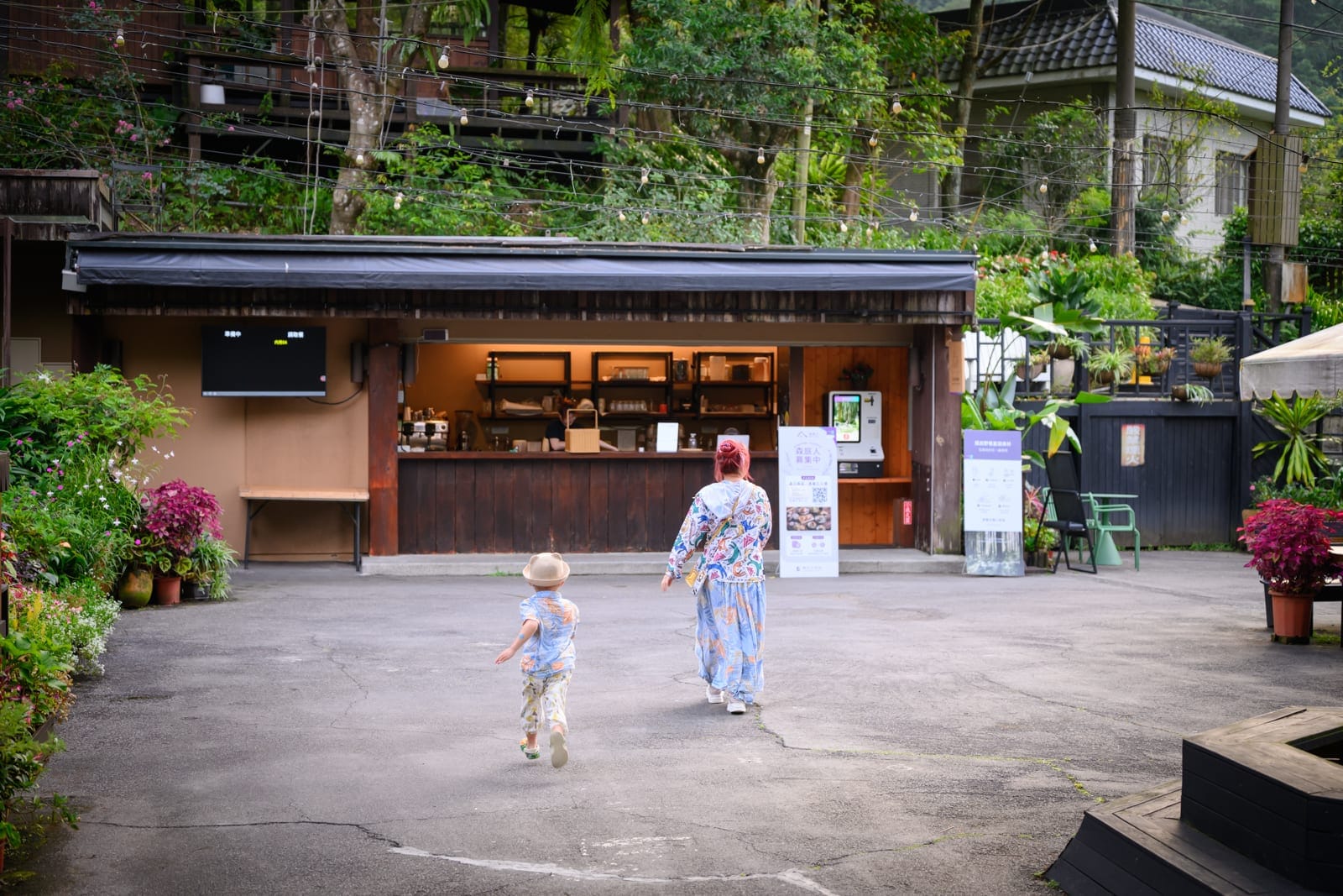 台中薰衣草森林住宿及園區｜廣場，櫃檯，山區建築，花圃，成年人，小孩。一名女子與戴帽兒童走在戶外灰色的空地，背對鏡頭朝向木造櫃檯