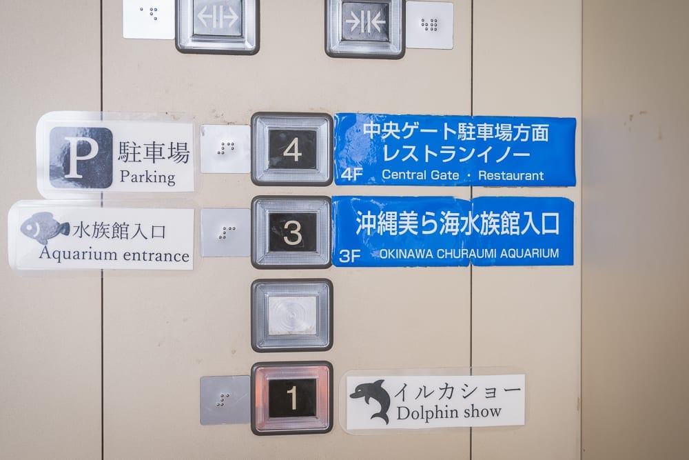 沖繩本部美麗海水族館完整攻略|巨型展缸、海豚池與親子戶外設施玩法解析 13 沖繩本部美麗海水族館|電梯按鈕、樓層面板、指引標示、日文標籤、無障礙點字。米色金屬牆面上排列金屬方框按鈕與數字標示,旁側貼